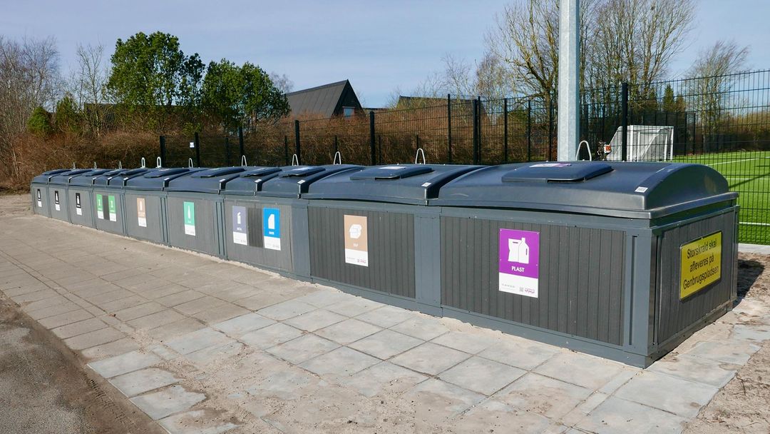 A long row of grey communal recycling containers at a bring site, each labelled for a different waste stream including glass, metal, paper, card and plastic