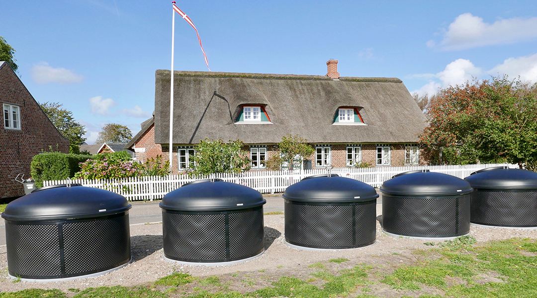 Five semi-buried waste containers with domed lids at a recycling point on Fan&oslash;, beside a thatched Danish house