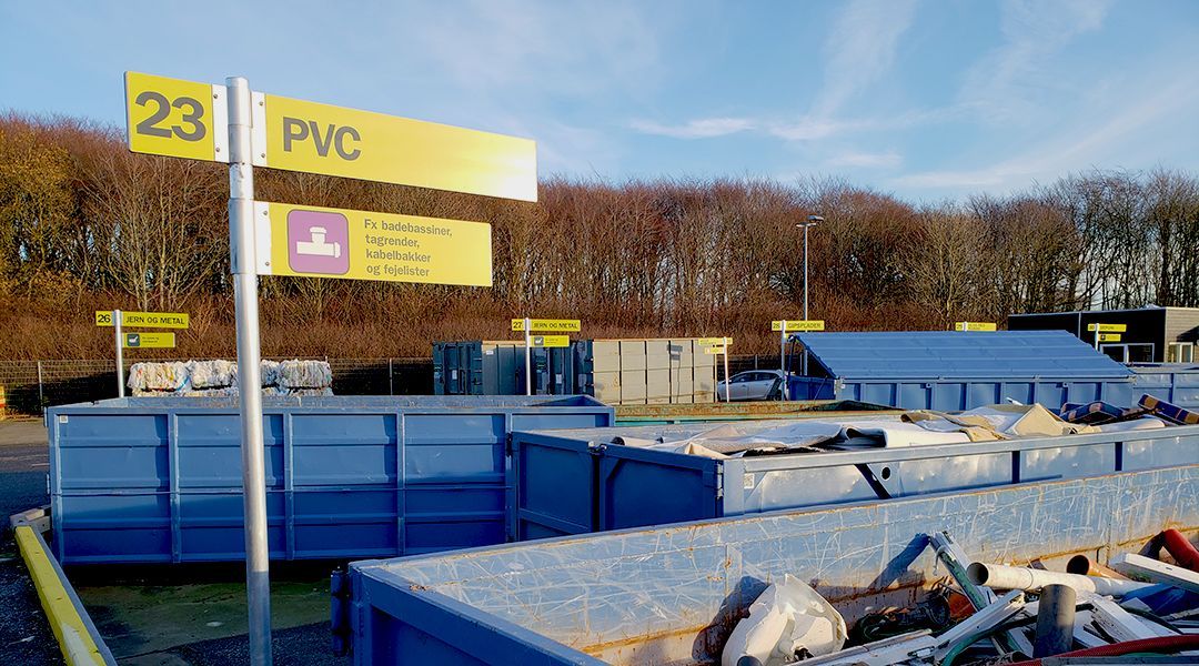 Open-top skips in numbered bays at a household waste recycling centre, with a yellow sign reading 23 PVC