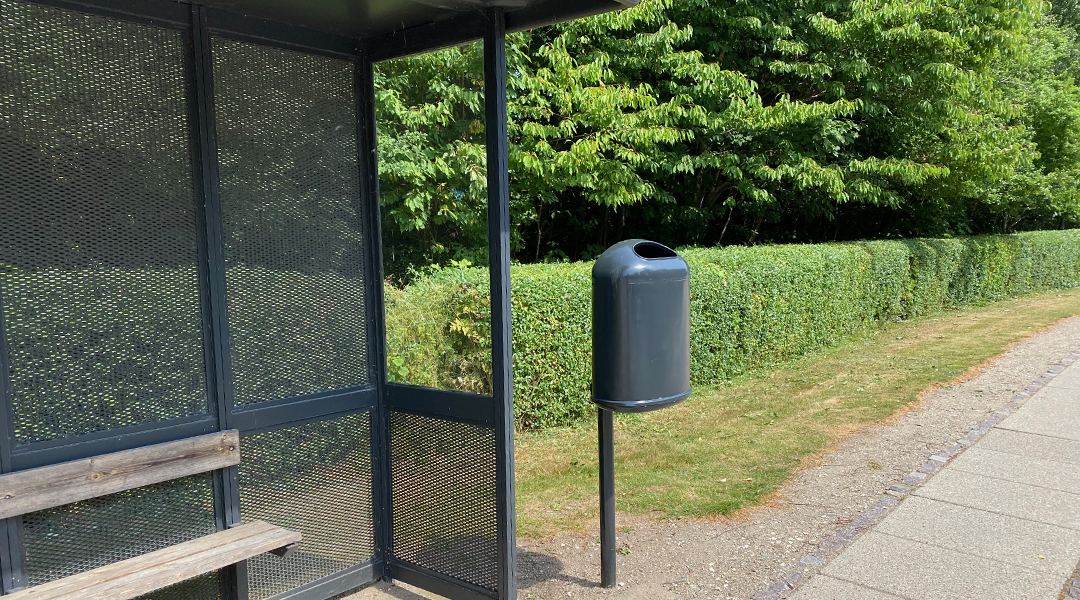 A public litter bin on a post beside a bus shelter, with a hedge and pavement behind